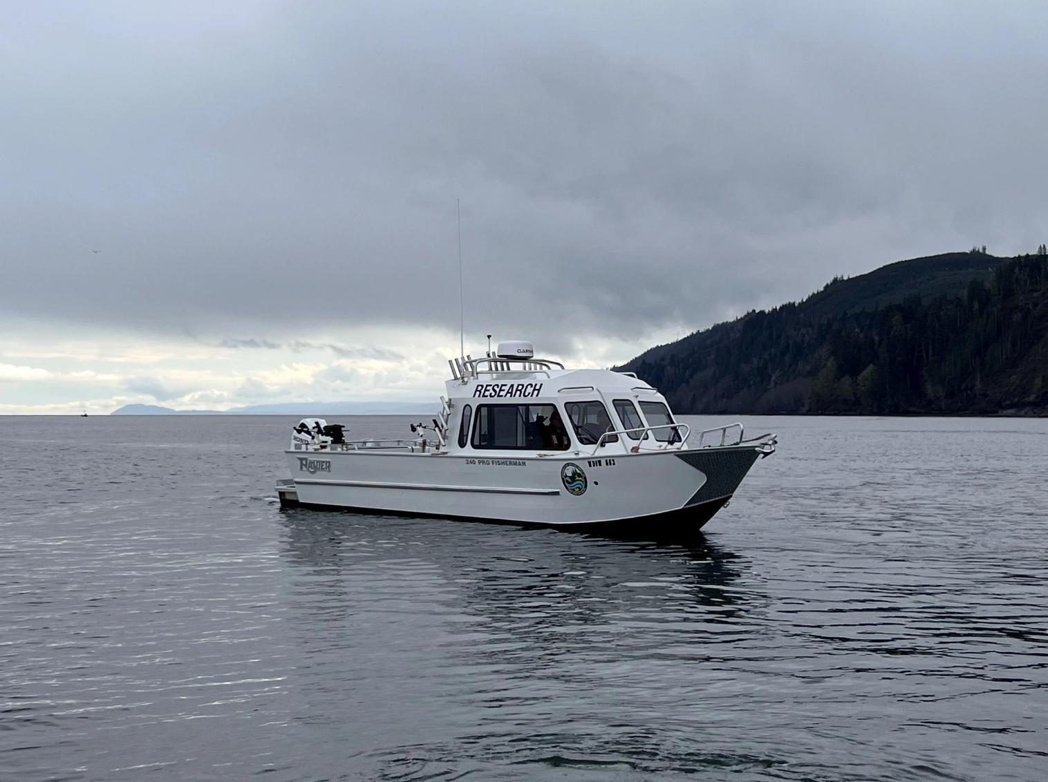 A white research vessel floats on calm water under a cloudy sky, with forested hills rising along the shoreline in the background. The boat is equipped with antennas and gear, and the word “RESEARCH” is visible on the cabin.